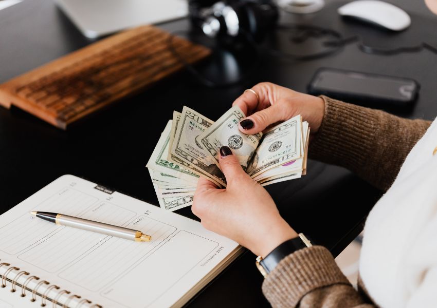 A landlord’s hands, pictured with black nail polish, count money over a desk with a notepad and pen.